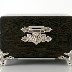 May include: A rectangular dark brown wooden box featuring a silver-toned metal latch with a shell-like design on the front. The box is supported by four silver-toned metal feet, each shaped like a bird with detailed wings and a beak.