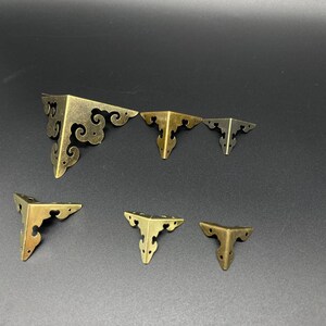 May include: Set of six antique gold corner brackets with ornate designs. The brackets are in various sizes and are perfect for adding a decorative touch to furniture or crafts.