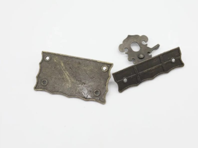 Op de afbeelding: Twee antieke metalen kastdeurgrepen. E&eacute;n greep heeft een decoratief ontwerp en de andere is een rechthoekige plaat.