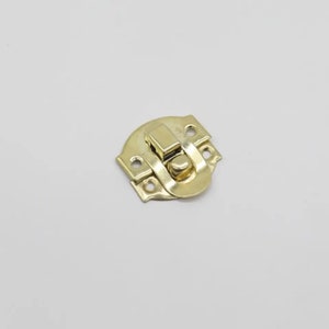 May include: A gold-toned metal latch with a rounded, decorative design. The latch has a small, round knob and a rectangular catch.