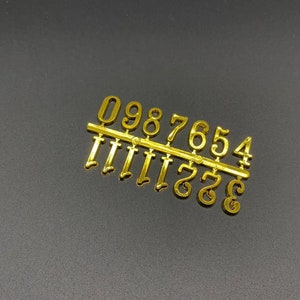 Puede incluir: Conjunto de números de plástico dorado con los números del 0 al 9. Los números están dispuestos en orden del 0 al 9, con el 0 a la izquierda y el 9 a la derecha.