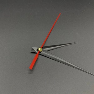 Puede incluir: Primer plano de una esfera de reloj con manecillas negras y un segundero rojo brillante. Las manecillas están posicionadas para indicar la hora. Las manecillas del reloj están unidas a un eje central de color latón. El fondo es gris oscuro liso.