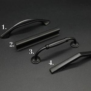 May include: Four black cabinet hardware pulls are displayed against a dark background. The pulls include a curved handle, a bar pull, a handle with a decorative center, and an angled handle. These are likely for kitchen or bathroom cabinets.