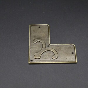 May include: An L-shaped metal corner bracket with an antique brass finish. The flat surface features a raised, curved decorative scroll pattern and three small circular holes positioned along the edges for mounting purposes.