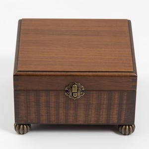 May include: A rectangular wooden box with a dark brown finish and a visible wood grain pattern. The box features a hinged lid, a central antique brass-colored metal latch on the front, and four small, rounded, decorative metal feet at the base.