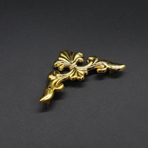 May include: Gold metal corner bracket with a decorative floral design. The bracket is shaped like a right angle and has a smooth, polished finish.