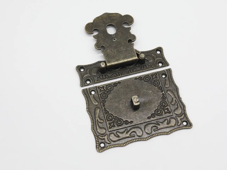 May include: A bronze-toned metal latch set consisting of two pieces. The top piece features a decorative cutout and hinge, while the bottom rectangular plate displays intricate embossed scrollwork patterns and a central protruding locking tab.