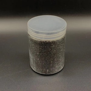 Pode incluir: Um frasco cilíndrico de plástico transparente preenchido com material granular preto fino. O recipiente possui uma tampa de rosca branca translúcida que está firmemente fixada ao topo do frasco, criando um recipiente de armazenamento simples e funcional.