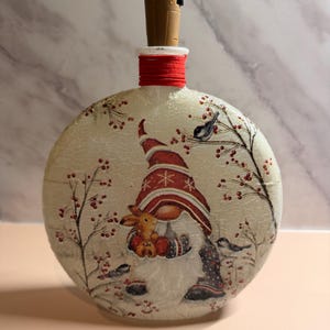 May include: A round, decorative bottle with a gnome design. The bottle features a gnome holding a small stuffed animal, surrounded by birds and berries. The bottle has a brown stopper and a red band around the neck.