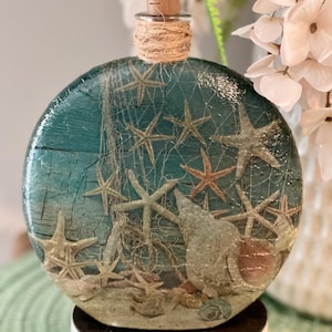 May include: A round, teal-colored bottle with a cork top and a twine accent. The bottle is decorated with a seashell and starfish design.