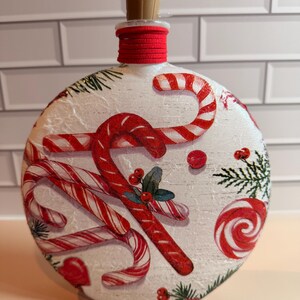 May include: A round, white, textured glass bottle featuring a festive holiday design of red and white candy canes, peppermint swirls, and green pine sprigs. The bottle neck is wrapped in red cord and topped with a tan cork stopper.