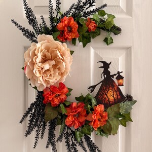 May include: A crescent-shaped wreath featuring a large pale peach peony, several smaller orange flowers, green ivy leaves, and black fern-like foliage. A silhouette cutout of a witch holding a lantern is attached to the right side of the wreath.