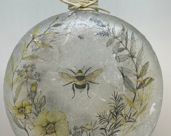May include: A round glass lamp base with a textured, white, and gray surface. The base features a floral design with a bee in the center. The lamp base is lit from within, creating a warm glow.