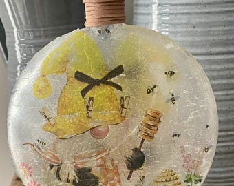 May include: A decorative, round glass bottle with a gnome and bee-themed design. The bottle features a yellow gnome hat, a jar of honey, and a beehive. Bees are scattered around the bottle. The bottle has a brown stopper and a light-colored base.