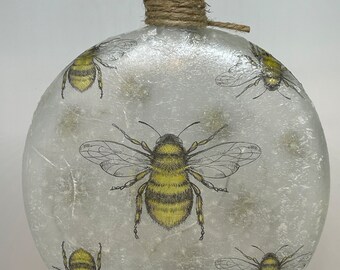 May include: A decorative glass bottle with a cork stopper and twine detail. The bottle is adorned with yellow and black bee illustrations. The bottle is round and has a frosted appearance.