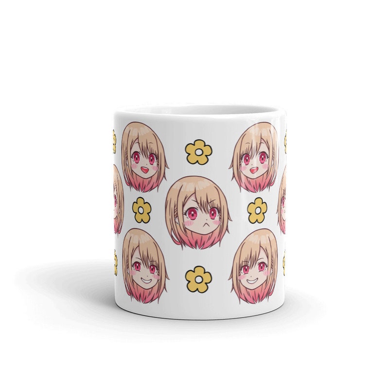 May include: White ceramic mug featuring a repeating pattern of a cartoon anime girl with blonde hair and pink cheeks. The girl has different facial expressions. The background is white with yellow flowers.