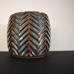 May include: A ceramic planter with a textured chevron pattern. The planter has a glossy finish in shades of blue and brown. The vessel is cylindrical and sits on a flat base. The design is a modern take on a classic shape.