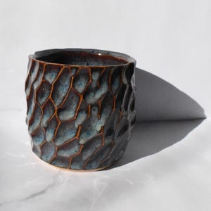 May include: A ceramic vessel with a textured, honeycomb design. The exterior has a dark, mottled glaze with blue and brown hues. The interior is a darker brown. This unique pottery piece would make a great addition to any home.