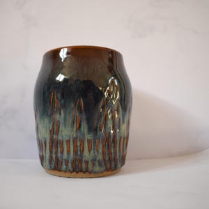 May include: A handmade ceramic vase with a glossy finish. The vase has a dark brown rim that fades into a deep blue-green body with vertical textured patterns in brown and tan. The vase has a wide opening and a solid base.