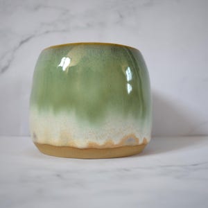 May include: A handcrafted ceramic pot with a rounded form. The exterior displays a colour gradient, shifting from light green at the top to creamy white at the base. The rim has a golden-yellow hue, and the base is a natural tan colour.