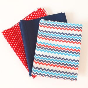 May include: A selection of fabric pieces in red, navy blue, and patterned designs. One fabric is red with white stars, another is solid navy blue, and the third features a chevron pattern in red, blue, and white. Suitable for sewing and craft projects.