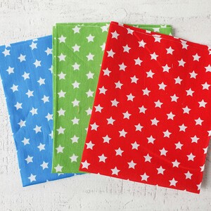 May include: Three folded fabric napkins in blue, green, and red, each patterned with white stars. The napkins are arranged on a white textured surface, showcasing the vibrant colours and design.