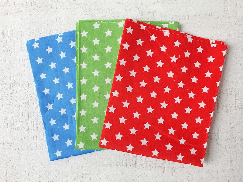 May include: Three folded fabric napkins in blue, green, and red, each patterned with white stars. The napkins are arranged on a white textured surface, showcasing the vibrant colours and design.
