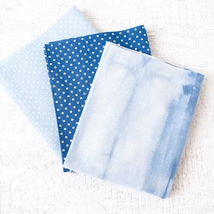 May include: Three folded fabric squares in shades of blue. One is a light blue with small white polka dots, another is a darker blue with white polka dots, and the third has a tie-dye effect. The fabrics are arranged on a white surface.