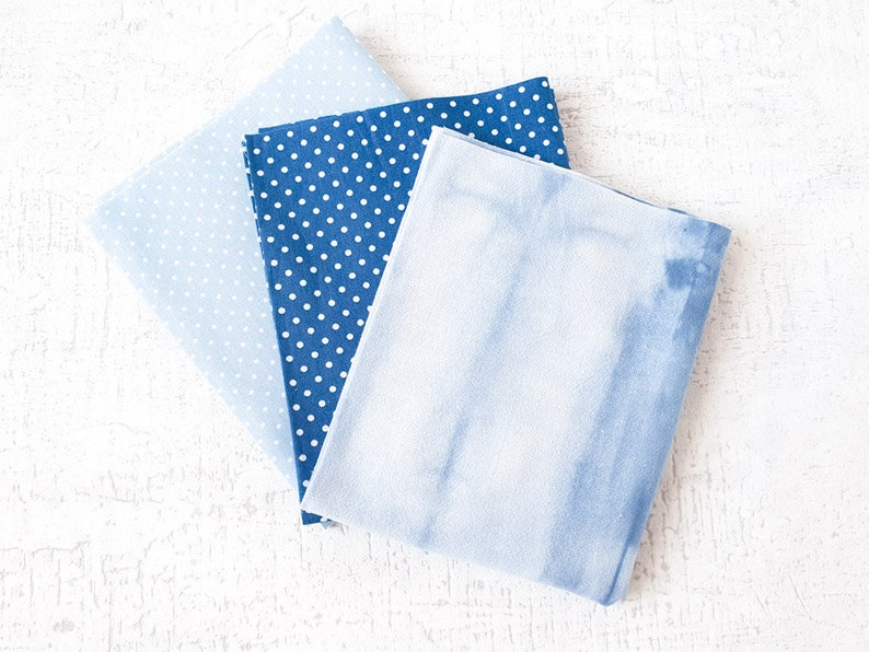 May include: Three folded fabric squares in shades of blue. One is a light blue with small white polka dots, another is a darker blue with white polka dots, and the third has a tie-dye effect. The fabrics are arranged on a white surface.