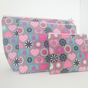 Op de afbeelding: Drie grijze ritszakjes met een roze, blauwe en zwarte bloemenprint. De zakjes zijn in elkaar genest, met het grootste zakje onderaan en het kleinste zakje bovenaan.