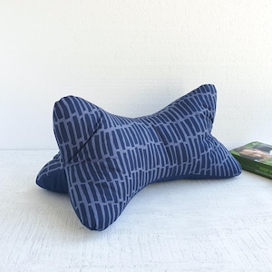 Può includere: Un cuscino blu navy a forma di osso con un motivo a righe verticali blu chiaro. Il cuscino è progettato per il supporto del collo. Un libro verde è visibile sullo sfondo.