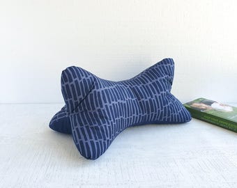 May include: A navy blue bone-shaped pillow with a light blue vertical stripe pattern. The pillow is designed for neck support. A green book is visible in the background.