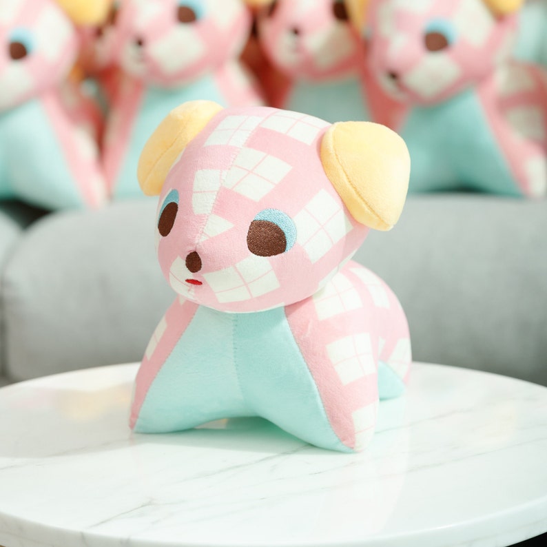 May include: A pink and blue plush toy dog with a checkered pattern. The dog has large brown eyes and yellow ears.