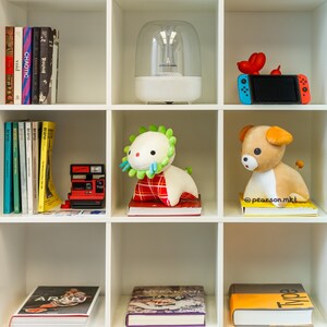 May include: A white shelf unit with six shelves. The top shelf has a white, round, dome-shaped object with a clear top. The second shelf has a blue and white Nintendo Switch console with a red and white controller. The third shelf has a plush toy with a green head and a red and white body. The fourth shelf has a plush toy with a brown head and a brown body. The fifth shelf has a red and black camera. The bottom shelf has three books with different covers.