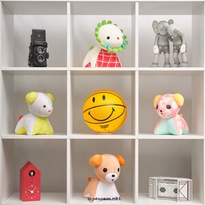 May include: A white shelf unit with nine compartments, each containing a different stuffed toy or object. The top row features a red and white plaid toy with a green crown, a black and white camera, and two gray and white figures. The middle row features a yellow smiley face ball, a white and yellow stuffed animal with a red heart, and a pink and white stuffed animal with a red bow. The bottom row features a red clock with white numbers, a brown and white stuffed animal with a pink nose, and a black and white box with a white label.
