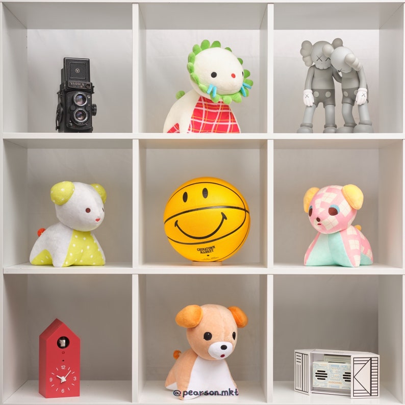 May include: A white shelf unit with nine compartments, each containing a different stuffed toy or object. The top row features a red and white plaid toy with a green crown, a black and white camera, and two gray and white figures. The middle row features a yellow smiley face ball, a white and yellow stuffed animal with a red heart, and a pink and white stuffed animal with a red bow. The bottom row features a red clock with white numbers, a brown and white stuffed animal with a pink nose, and a black and white box with a white label.