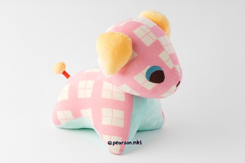 May include: A pink and white checkered plush toy dog with yellow ears and blue eyes. The dog is sitting on a white surface. The toy is made of soft, cuddly fabric and is perfect for cuddling.