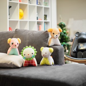 May include: Four plush toys, including a pink and blue bear, a green and red creature, a yellow and white bear, and a brown and white bear, are sitting on a gray couch. The toys are all facing the camera. The couch is in a living room with a bookshelf and a wooden chair in the background.