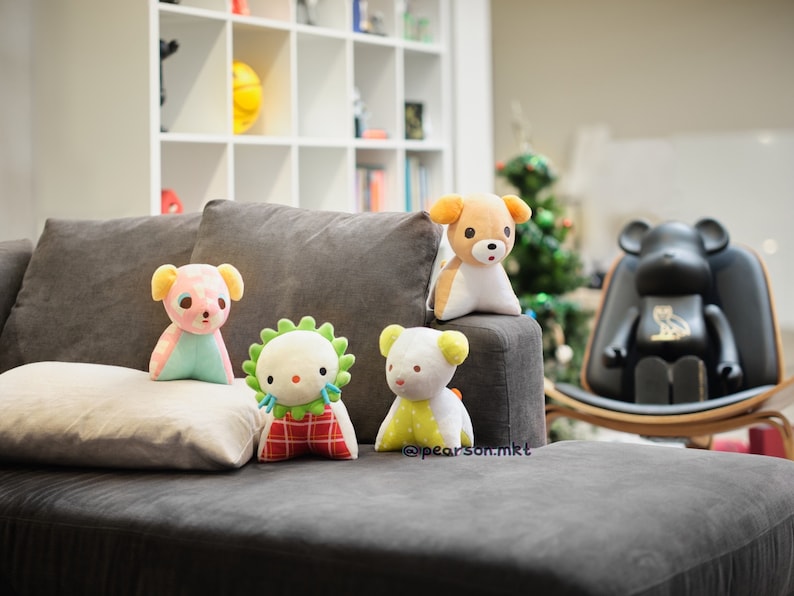 May include: Four plush toys, including a pink and blue bear, a green and red creature, a yellow and white bear, and a brown and white bear, are sitting on a gray couch. The toys are all facing the camera. The couch is in a living room with a bookshelf and a wooden chair in the background.