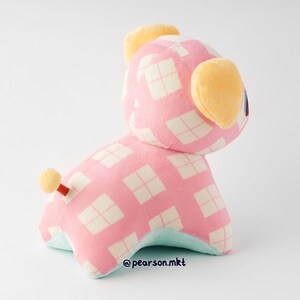 May include: Pink and white checkered plush toy with yellow ears and a small tail. The toy is sitting on a white surface.