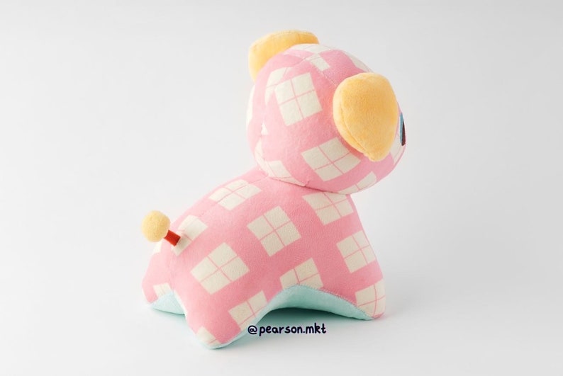 May include: Pink and white checkered plush toy with yellow ears and a small tail. The toy is sitting on a white surface.
