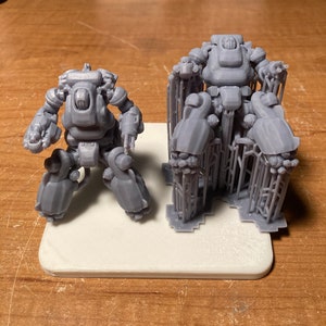 Pode incluir: Dois robôs cinzentos impressos em 3D, um em pé e outro sentado, em uma plataforma branca. O robô em pé tem vários braços e pernas, enquanto o robô sentado tem dois braços e duas pernas.