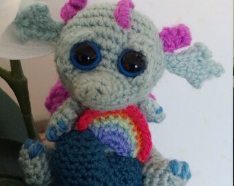 May include: A small crocheted dragon toy with blue eyes, pink ears, and a rainbow crocheted heart. The dragon is sitting on a gray pot with rocks.