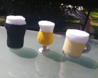 May include: Three crocheted beer glasses, one black, one yellow, and one with a white top and a yellow bottom. The glasses are sitting on a glass table.
