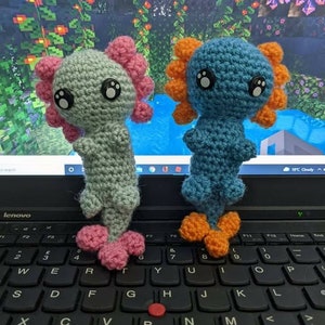 May include: Two crocheted axolotl plushies, one light blue with orange fins and one light green with pink fins, sitting on a black laptop keyboard.