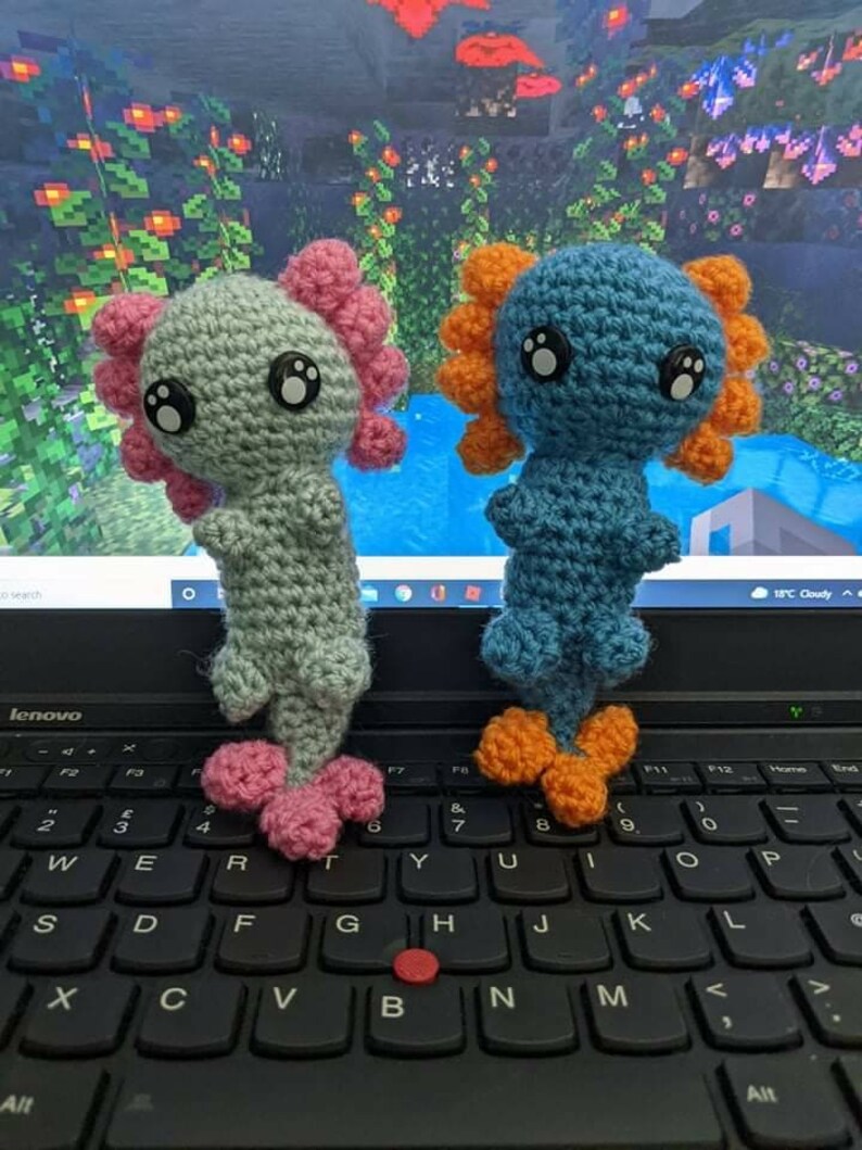 May include: Two crocheted axolotl plushies, one light blue with orange fins and one light green with pink fins, sitting on a black laptop keyboard.