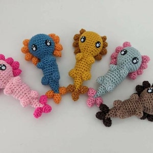 May include: Five crocheted axolotl plushies in different colors. The axolotls are pink, blue, brown, yellow, and light blue. Each axolotl has large black eyes and a small mouth.