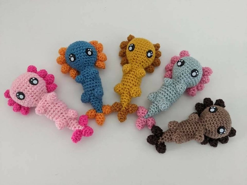 May include: Five crocheted axolotl plushies in different colors. The axolotls are pink, blue, brown, yellow, and light blue. Each axolotl has large black eyes and a small mouth.
