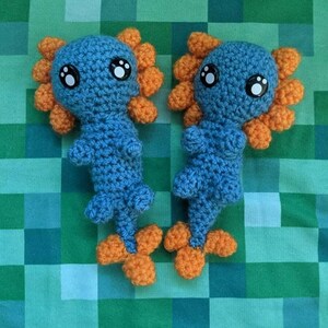 May include: Two crocheted axolotl plushies with blue bodies, orange frills, and black eyes. The plushies are sitting on a green and blue checkered background.