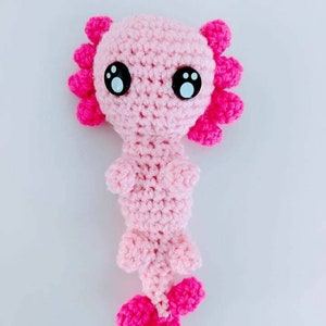 May include: A pink crocheted axolotl plush toy with black eyes. The axolotl has pink frills around its head and a pink tail.